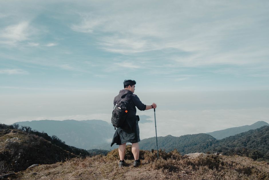 Solo hiker with backpack and walking stick looking at breathtaking mountain landscape under blue sky.